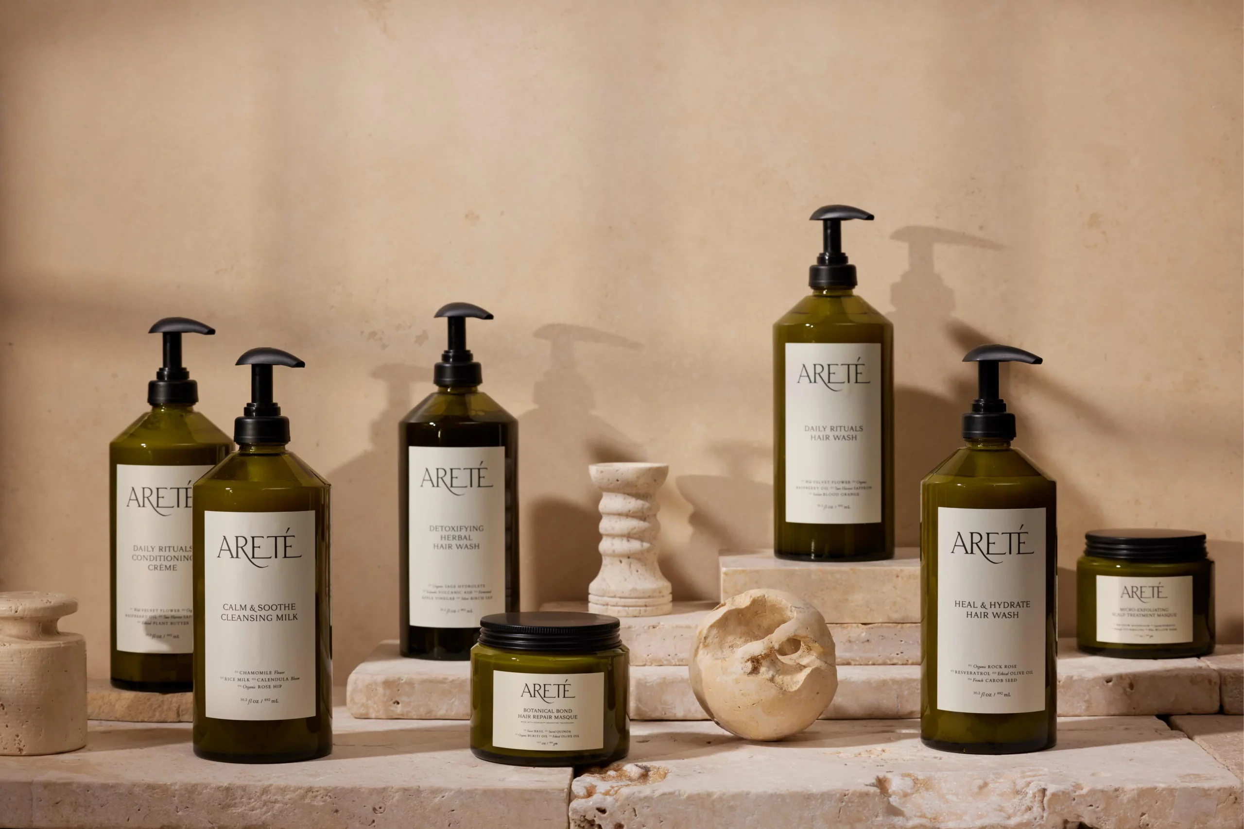 A collection of Areté skincare and haircare products arranged on beige stone blocks, including bottles of hair wash, conditioning crème, cleansing milk, and a hair repair masque in green packaging with black pumps.